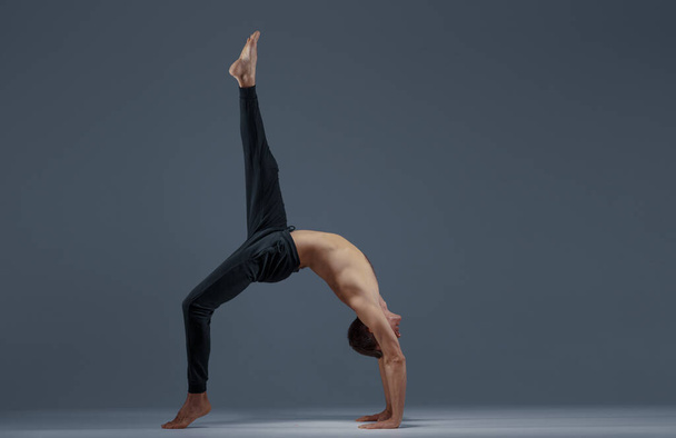 Male yoga doing stretching exercise in studio, grey background. Strong man practicing yogi , asana training, top concentration, healthy lifestyle - Valokuva, kuva