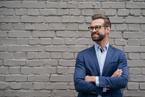 Porträt eines selbstbewusst lächelnden Managers mit Businessanzug und stylischer Brille auf grauem Hintergrund. Schöner Geschäftsmann mit verschränkten Armen, der den Kopierraum anschaut - Foto, Bild