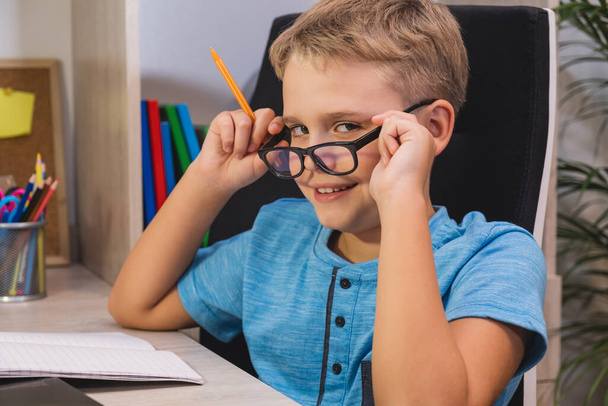 Home learning, online education. Schoolboy with glasses studies at home. Boy while sitting at table in front of laptop and looks at camera. - Фото, изображение