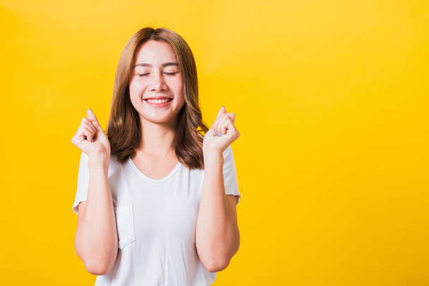 Asian Thai happy portrait beautiful cute young woman standing wear t-shirt makes raised fists up celebrating her winning success she close eyes isolated, studio shot on yellow background with copy space - Photo, image