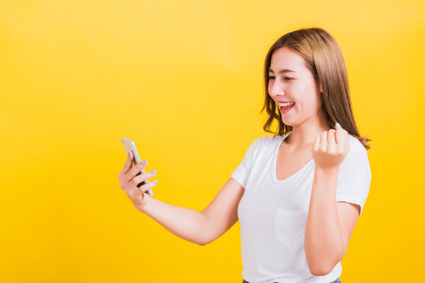 Asian Thai portret szczęśliwy piękny ładny młoda kobieta uśmiecha się nosić t-shirt podnoszenia ręki świętując zwycięstwo z inteligentnym telefonem komórkowym patrząc na telefon odizolowany, na żółtym tle z miejsca kopiowania - Zdjęcie, obraz