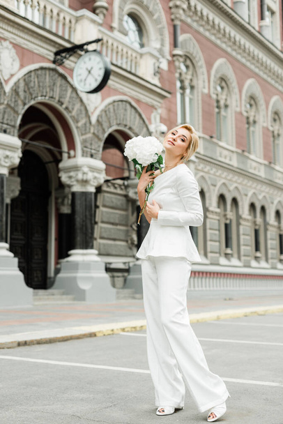 Mladá stylová dáma v bílém kalhotovém kostýmu s elegantním účesem a kyticí hortenzických květin na ulicích města. Ta žena je nevěsta. Měkké selektivní zaměření - Fotografie, Obrázek