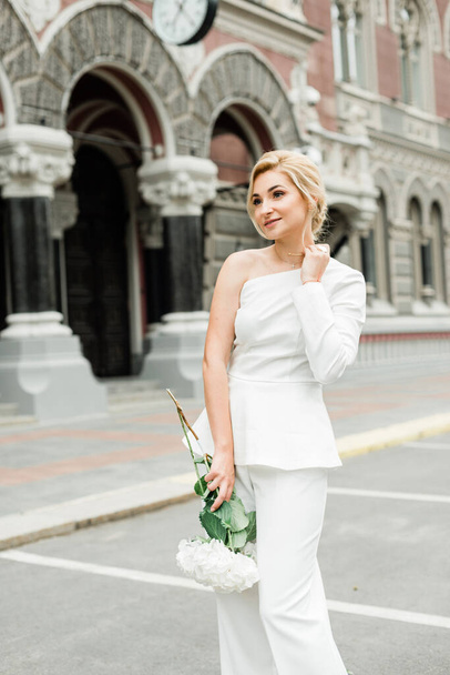 A young stylish lady in a white pantsuit with an elegant hairstyle with a bouquet of hydrangea flowers on the streets of the city. The woman is the bride. Soft selective focus - Фото, изображение