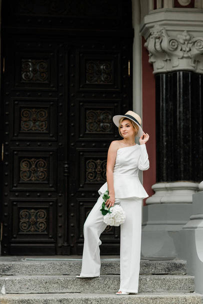 Νεαρή κομψή κυρία με λευκό κοστούμι παντελονιού και καπέλο με κομψό χτένισμα με μπουκέτο λουλούδια ορτανσίας στους δρόμους της πόλης. Η γυναίκα είναι η νύφη.. - Φωτογραφία, εικόνα