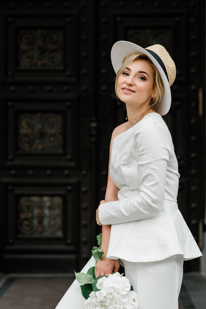 Mladá stylová dáma v bílém kalhotovém kostýmu a klobouku s elegantním účesem s kyticí květin hortenzie na ulicích města. Ta žena je nevěsta.. - Fotografie, Obrázek
