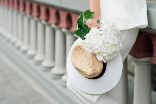 白いスーツを着たスタイリッシュな女性と街の路上でアジサイの花束と帽子。柔らかい選択的フォーカス - 写真・画像