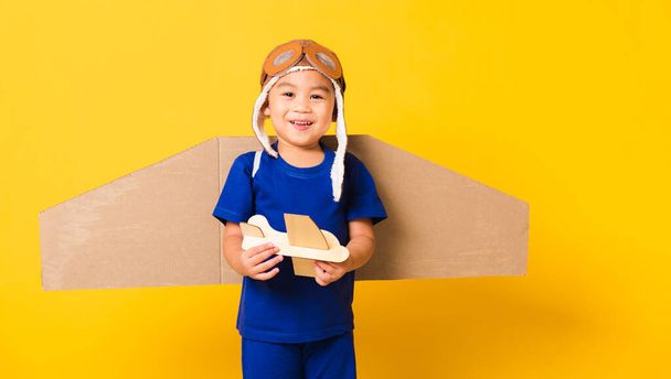 Happy Asian handsome funny child or kid little boy smile wear pilot hat play and goggles with toy cardboard airplane wings fly hold plane toy, studio shot isolated yellow background, Startup freedom - Foto, Imagem