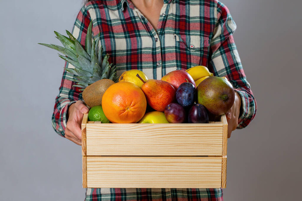 Person holding wooden crate filled with fresh colorful fruit. Woman in flannel shirt delivering wooden box full of ripe sweet food from front. Female figure with healthy juicy plants. - Foto, imagen