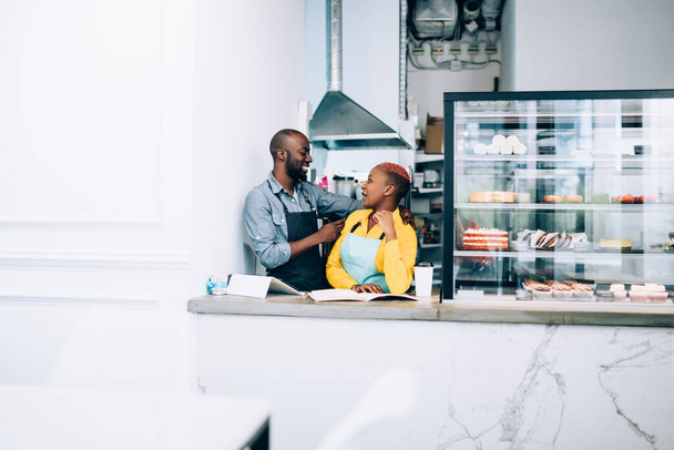 Cheerful African American business partners looking at each other and laughing while standing beside counter and showcase with desserts in light modern pastry shop - 写真・画像