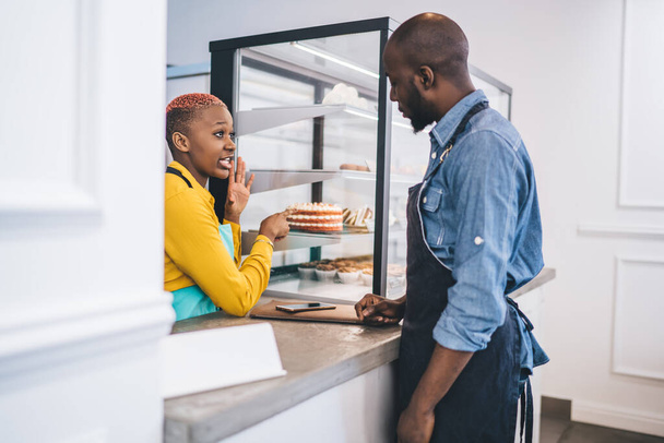 Side view of young African American man in uniform waiting for cake and speaking with stylish black woman in yellow shirt and turquoise apron suggesting quantity of pieces  - Foto, Imagem