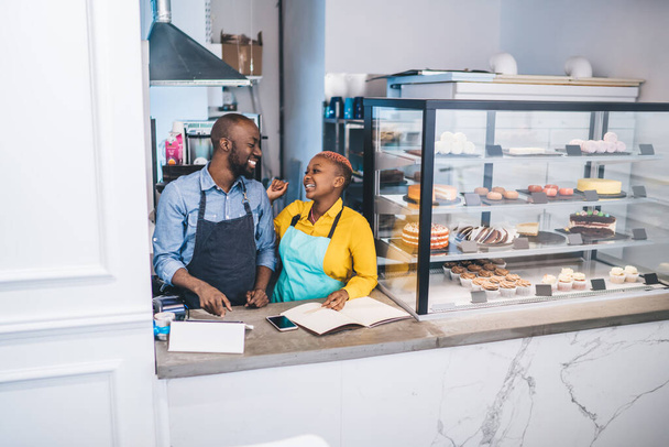 Stylish African American woman in yellow shirt and turquoise apron touching black man in dark apron and laughing at country of pastry shop - 写真・画像