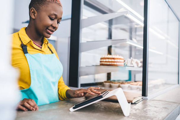 Focused lovely African American young female with short hairstyle in apron doing work using tablet standing at counter in bakery - Foto, Imagem