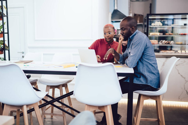 Skoncentrowany inteligentny Afroamerykanin współczesnych młodych współpracowników przeglądających na laptopie siedzi przy stole z licznikiem w tle w kawiarni w mieście - Zdjęcie, obraz