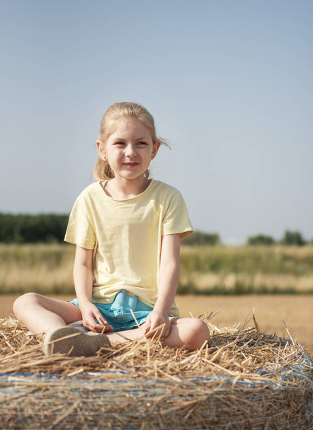 Küçük kız yaz günü buğday tarlasında eğleniyor. Hasat zamanı saman balyasında oynayan çocuk. - Fotoğraf, Görsel