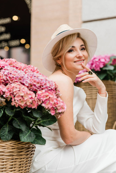 ピンクのアジサイの花の間で白いジャケットと帽子の美しいエレガントな女性の肖像画。柔らかい選択的フォーカス. - 写真・画像
