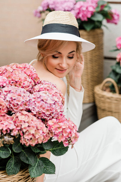 Porträt einer schönen eleganten Frau in weißer Jacke und Hut inmitten rosa Hortensien. Weiche selektive Fokussierung. - Foto, Bild