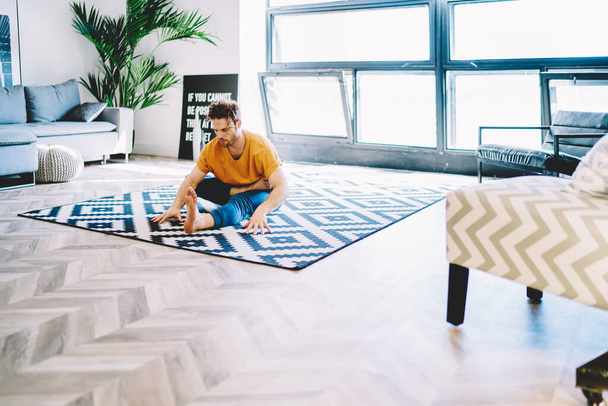 Fit casual man doing yoga asana on colorful carpet in modern living room with big windows and plant in pot - Fotografie, Obrázek