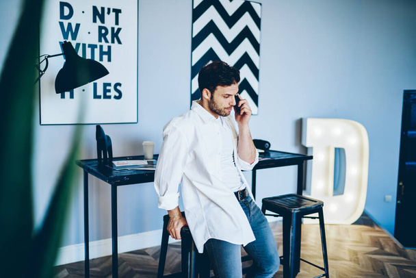 Anxious male in white shirt and white t shirt making call on smartphone leaning on chair in creative workplace with black and white picture on wal - Fotoğraf, Görsel