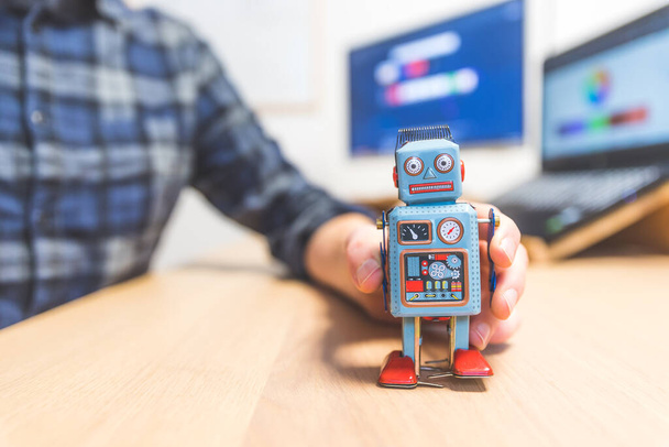 Toy robot on a desktop, man working on the computer in the background. Symbol for artificial intelligence, chatbots or social bots and algorithms - Fotoğraf, Görsel