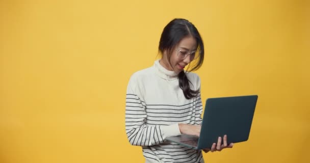 Gelukkig aantrekkelijk Azië freelancer dame met casual doek houden laptop computer en genieten van werken dans zingen en typen werk op kopieerruimte terwijl geïsoleerd over gele achtergrond. - Video