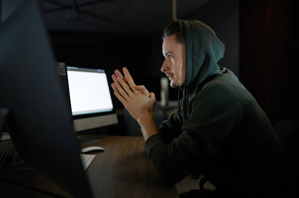 Young male internet hacker in hood sitting at monitors. Illegal web programmer at workplace, criminal occupation. Data hacking, cyber security - Фото, зображення