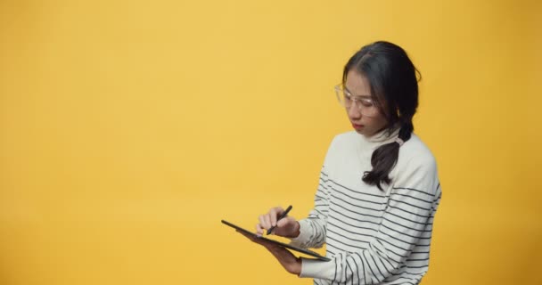 Happy attractive Asia student lady holding tablet computer and pointing finger up at copy space enjoy looking book digital online shopping while standing isolated over yellow background. - Footage, Video