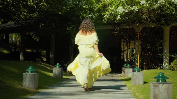 A young European girl walking in the backyard with a green garden and grass in a beautiful long yellow dress, turning her back - Foto, Bild
