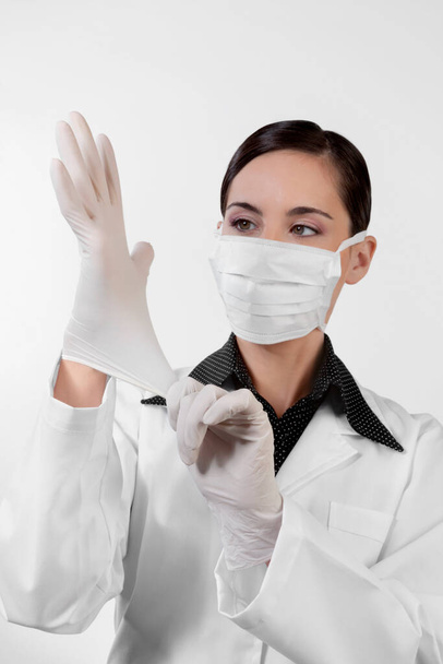 young woman in a white coat and gloves is holding a syringe and a mask on a gray background - Foto, Imagem
