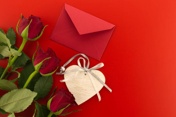 Red roses, envelope and wooden heart medallion on red background with free copy space. Wishes, greetings and love message - Fotó, kép