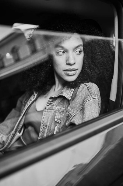 Czarno-biały portret z odbiciami pięknej Afroamerykanki czarnowłosej kierowcy siedzącej w nowym samochodzie, cieszącej się podróżą. Kursy jazdy i koncepcja ubezpieczenia na życie - Zdjęcie, obraz