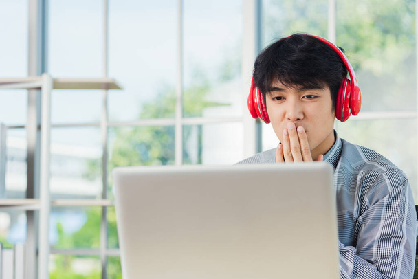 Азиатский молодой бизнесмен счастье с красными наушниками он сидит на рабочем столе домашний офис ищет ноутбук монитор компьютера, уверенный красивый мужчина образ жизни улыбка расслабляет он слушает музыку - Фото, изображение