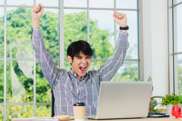 Yes! Happy excited Asian man raising his arm up to celebrate celebrating success. Young businessman using laptop computer at office desk he glad to receive good winner profits from the job - Photo, Image