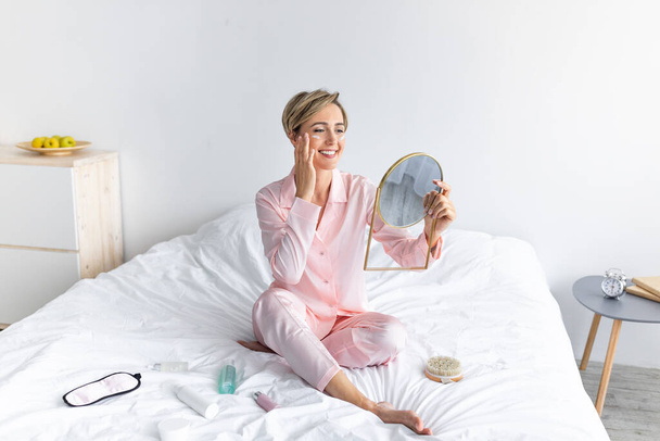 Portrait of smiling woman holding mirror applying cream on cheeks - Foto, imagen