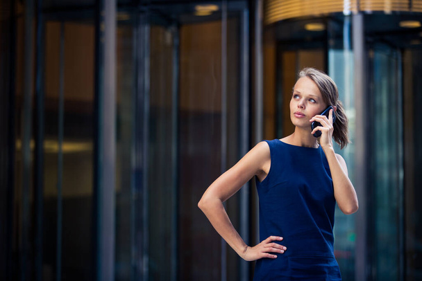 Gorgeous young businesswoman calling on her phone in front of her company's building (shallow DOF  color toned image) - Foto, afbeelding