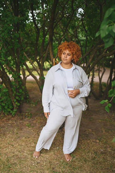Modeporträt einer jungen, natürlich schönen Afroamerikanerin in hellem Hosenanzug mit afroroten Haaren, die in einem Naturpark in grünem Laub posiert. Diversität und körperliches positives Konzept. - Foto, Bild
