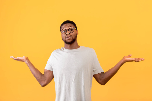 Confused black guy raising hands up and looking at camera, standing over yellow studio background - Photo, Image