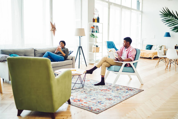 Young African American couple in casual clothing communicating and browsing devices in modern living room with big windows on sunny day  - Fotoğraf, Görsel