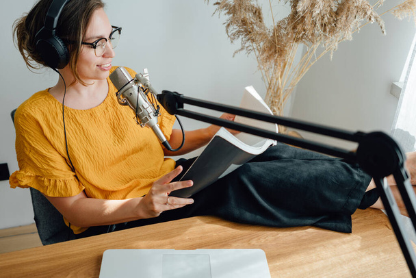 Woman podcaster streaming and broadcasting her podcast from homemade studio. Female recording podcast from home and talking into a microphone. - Fotografie, Obrázek