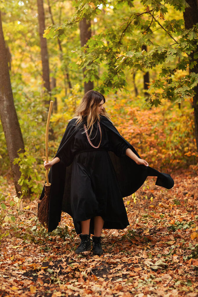 Блондинка в костюме ведьмы с метлой идущей по тропинке покрытой осенними листьями в день Хэллоуина в лесу - Фото, изображение