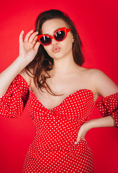 Stijlvolle aantrekkelijke jonge vrouw poseren in rode polka dot nauwsluitende jurk en in kat bril, staan in flirten pose met lippen gevouwen in kus, geïsoleerd over rode studio achtergrond - Foto, afbeelding