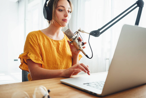 Female podcaster recording her podcast using microphone and laptop at his home broadcast studio. Woman streaming live from homemade radio studio - 写真・画像