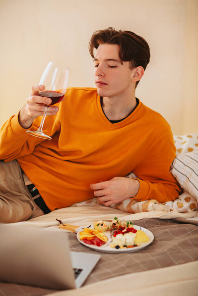 Χαλαρός νεαρός με ένα ποτήρι κρασί στα χέρια του ξαπλωμένος στο άνετο κρεβάτι με ένα πιάτο φαγητό, βλέποντας βίντεο στο λάπτοπ ενώ τρώει, απολαμβάνοντας τον ελεύθερο χρόνο του χαλαρώνοντας στο σπίτι - Φωτογραφία, εικόνα