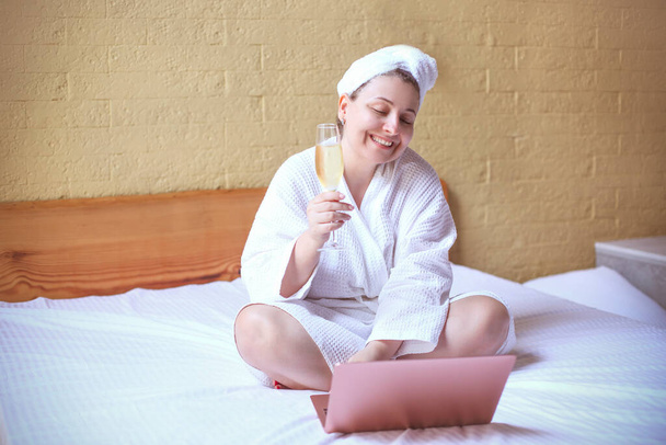 Jeune femme hipster souriant en buvant du champagne lors d'un appel vidéo via un ordinateur portable à la maison assis sur le lit - Photo, image