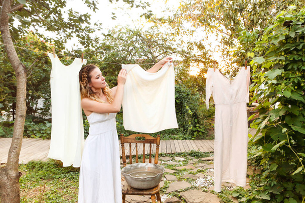 Młoda kobieta wieszająca pranie na zewnątrz. Cute girl w sukience mycia białych ubrań w metalowej umywalce na podwórku, wiszące pranie na sznurze i pozostawiając go do wyschnięcia w ogrodzie, suszenia ubrań na zewnątrz na wietrze - Zdjęcie, obraz
