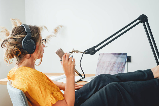 Young woman recording podcast on his laptop computer with headphones and  microphone. Female podcaster streaming her voice into mike at home studi - Zdjęcie, obraz