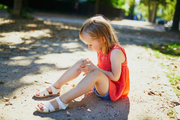 Cute little girl sitting on the ground after falling down. Child getting hurt while walking in park. Kid looking at her boo-boo - Φωτογραφία, εικόνα