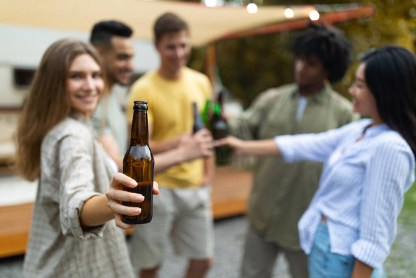 Bonita dama caucásica con sus diversos amigos brindando con botellas de cerveza, bebiendo alcohol cerca de RV al aire libre en otoño - Foto, Imagen