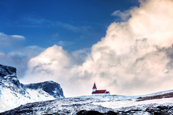 Vik i Myrdal Kirche, in Vik, Island, vor verschneiten Bergen und blauem Himmel mit rollenden Wolken. Diese malerische, rot gedeckte Kirche thront auf einem Hügel über dem Dorf im Süden Islands. - Foto, Bild