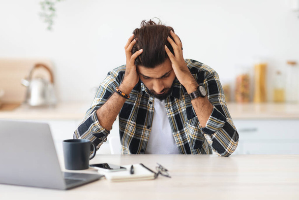 Депрессивный мужчина, сидящий перед ноутбуком и трогающий свою голову - Фото, изображение