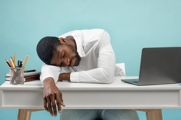 Junge erschöpfte schwarze Angestellte schläft am Arbeitsplatz, ist der Arbeit überdrüssig und legt den Kopf auf den Tisch, blauer Hintergrund - Foto, Bild